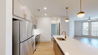 This single-floor residence showcases a thoughtfully designed open-concept layout that seamlessly connects the kitchen and living areas. The kitchen serves as a centerpiece of the home, anchored by a large island with a white quartz countertop, an undermount sink, and a brushed gold faucet. Overhead, two globe pendant lights with gold hardware add a warm, contemporary touch. The kitchen is equipped with a full suite of stainless steel appliances — including a refrigerator, range, and built-in microwave — complemented by taupe shaker-style cabinetry with gold hardware and a clean white tile backsplash. Light wood-look flooring runs throughout the main living areas, lending warmth and continuity to the space. The living area is generously proportioned and benefits from natural light through French doors that open to a balcony, with a ceiling fan providing additional comfort. The bedroom features plush carpet flooring, a ceiling fan, and a window with natural light, while an adjacent walk-in closet provides ample storage with wire shelving on multiple walls. The bathroom is finished with tile flooring, a full tub-shower combination with sliding glass doors, and chrome fixtures, completing this well-appointed residence.