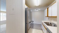 Located at 2240 Skillman Ave E in North St. Paul, Minnesota, this one-bedroom unit offers a practical and well-organized single-floor layout. The galley-style kitchen is efficiently arranged, featuring stainless steel appliances including a refrigerator and range, white cabinetry with wood-tone trim, a double stainless steel sink with white countertops, and a dishwasher. Light tile flooring runs throughout the kitchen, providing easy maintenance. A hallway connects the kitchen to the main living area, which transitions to soft, neutral-toned carpet. The open living space is bathed in natural light from a large window fitted with blinds, and includes a baseboard heating system along the exterior wall, ensuring comfort during Minnesota winters. The textured ceiling and warm beige walls carry throughout the unit, creating a cohesive and understated aesthetic. The bedroom offers a comfortable footprint with plush carpeting, a sliding-door closet, baseboard heat, and its own window with outdoor views of the surrounding neighborhood. The overall layout is straightforward and functional, making efficient use of space throughout.