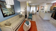 This single-story space showcases an open-concept floor plan that connects the living, dining, and kitchen areas in a fluid, functional layout. The living area serves as the focal point, featuring plush carpeting, crown molding, and a warm, inviting atmosphere. A teal accent wall adds a bold pop of color, complementing the neutral beige tones found throughout the space. The living room comfortably accommodates a full-sized sofa, accent chair, and oval coffee table, making it well-suited for both relaxing and entertaining.

Moving through the space, the layout transitions naturally into a dedicated dining area, which benefits from abundant natural light streaming through large windows dressed with curtains. The dining space is furnished with a round table and upholstered chairs, creating an elegant yet casual setting. Adjacent to the dining area, the kitchen is partially visible and features pendant lighting and a countertop bar, providing additional seating and convenience. The consistent use of carpet and crown molding throughout ties the entire space together, delivering a cohesive and well-appointed living environment.