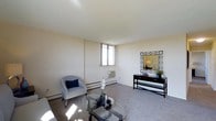 This single-floor space offers a clean, functional layout with a comfortable living area as its centerpiece. The living room features neutral wall-to-wall carpeting and cream-colored walls that create a bright, airy atmosphere. Natural light enters through a window positioned along one wall, which also houses a wall-mounted air conditioning unit. Baseboard heating runs along the perimeter, ensuring year-round climate comfort. The room is well-proportioned, providing ample space for seating arrangements and accent furniture, as demonstrated by the glass-top coffee table, upholstered accent chair, and a sleek console table topped with a geometric-framed mirror.

One of the most notable features of this space is its elevated position, which affords expansive panoramic views from the balcony. Looking out, residents are treated to sweeping sightlines over a lush urban tree canopy, surrounding residential neighborhoods, commercial buildings, and open sky stretching to the horizon — a striking contrast to the understated interior. The outdoor vantage point captures both the density of the surrounding city and the greenery woven throughout it, making the views a defining characteristic of this space.
