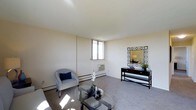 This single-floor space offers a clean, functional layout with a comfortable living area as its centerpiece. The living room features neutral wall-to-wall carpeting and cream-colored walls that create a bright, airy atmosphere. Natural light enters through a window positioned along one wall, which also houses a wall-mounted air conditioning unit. Baseboard heating runs along the perimeter, ensuring year-round climate comfort. The room is well-proportioned, providing ample space for seating arrangements and accent furniture, as demonstrated by the glass-top coffee table, upholstered accent chair, and a sleek console table topped with a geometric-framed mirror.

One of the most notable features of this space is its elevated position, which affords expansive panoramic views from the balcony. Looking out, residents are treated to sweeping sightlines over a lush urban tree canopy, surrounding residential neighborhoods, commercial buildings, and open sky stretching to the horizon — a striking contrast to the understated interior. The outdoor vantage point captures both the density of the surrounding city and the greenery woven throughout it, making the views a defining characteristic of this space.