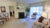 This single-floor residence offers a thoughtfully designed layout with a refined, classic aesthetic. The living room serves as the centerpiece of the home, anchored by a sleek fireplace with a decorative mantle adorned with a framed mirror and accent pieces, adding a touch of sophistication. Soft, neutral-toned carpeting extends throughout the space, contributing to the warm and comfortable atmosphere.

Large sliding glass doors and sheer curtains allow an abundance of natural light to flood the living area, offering views of lush greenery just beyond the exterior. A tall, potted palm plant placed beside the doors reinforces a fresh, natural element within the interior. The living room is furnished with a glass-top coffee table, elegant upholstered seating, and accent chairs with decorative cushions, creating a cohesive and inviting arrangement.

Crown molding and recessed lighting add architectural detail to the ceilings, elevating the overall finish of the space. Passageways visible from the main living area suggest a well-connected floor plan with additional rooms accessible nearby. Overall, the space presents a bright, clean, and tastefully styled interior suited for comfortable everyday living.