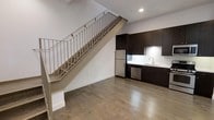 This two-story space offers a clean, modern aesthetic defined by an open-concept layout and thoughtful design throughout. The main level features warm hardwood flooring that extends across the living and kitchen areas, creating a cohesive and inviting atmosphere. A central staircase with a contemporary metal railing serves as a striking architectural element, connecting the two floors while adding visual interest to the space.

The kitchen is outfitted with dark espresso cabinetry that provides a bold contrast against the crisp white walls and countertops. Stainless steel appliances, including a refrigerator and dishwasher, lend a polished, professional quality to the kitchen. A stainless steel sink with a modern faucet completes the setup, making this kitchen as functional as it is stylish.

Recessed lighting illuminates the main level, enhancing the bright and airy feel of the space. The white walls and high ceilings amplify the sense of openness, allowing light to flow freely throughout. The second floor offers additional living space accessible via the interior staircase. Overall, this space combines contemporary finishes with a practical layout, making it a versatile and well-appointed environment.
