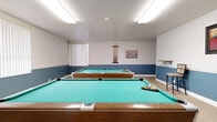This dedicated billiards room is a well-appointed recreational space designed for both casual play and social entertainment. The room comfortably accommodates two full-size pool tables with teal felt surfaces and classic wood-and-metal frames, positioned to allow players ample room to maneuver on all sides. The interior is finished with a distinctive two-tone wall treatment, featuring a calming steel blue lower half contrasted against crisp white upper walls, giving the space a clean and inviting character. Neutral carpeted flooring runs throughout, contributing to a comfortable and low-maintenance environment. Overhead, flush-mounted fluorescent light fixtures provide bright, even illumination across the playing surfaces. Multiple vertical blind-covered windows line the walls, allowing natural light to filter into the room while maintaining privacy. A wall-mounted cue rack keeps billiard cues neatly organized and readily accessible. Decorative framed artwork adds a modest aesthetic touch to the walls, while small wall-mounted shelves offer convenient space for personal items. A round wall clock and a compact chair are also present, rounding out the functional furnishings of the space. The room is accessed through a standard interior door, making it a self-contained amenity well-suited for community or shared-use purposes.