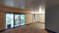 This single-floor space presents a straightforward and functional layout centered around an open living area. The room is finished with neutral-toned walls and carpeted flooring, creating a simple and adaptable interior. A prominent set of sliding glass doors dominates one wall, opening onto a balcony that frames views of mature, verdant trees — bringing a sense of the outdoors in and filling the space with natural light. Adjacent to the sliding doors, a window with light curtains provides an additional source of daylight, enhancing the airy feel of the room. The ceiling is smooth and flat, contributing to the clean, uncluttered aesthetic of the space. Dark baseboards run along the perimeter of the room, offering a subtle contrast to the lighter walls. A built-in shelving nook near the window adds a practical storage element. The overall ambiance is calm and neutral, offering a blank canvas well-suited for a variety of uses. The connection to the outdoors through both the balcony and the window views is a defining characteristic of this space, making natural scenery a central part of the interior experience.