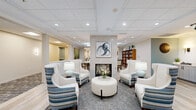 Located at 1470 S Havana St in Aurora, Colorado, this thoughtfully designed communal space offers a warm and inviting atmosphere across a single, well-appointed floor. The layout seamlessly integrates several distinct functional zones, each contributing to an overall sense of comfort and community.

At the heart of the lounge area, a floor-to-ceiling tiled fireplace serves as a striking focal point, flanked by elegant wingback chairs upholstered in crisp white and accented with blue-striped fabric. A circular tufted ottoman anchors the seating arrangement, while recessed lighting and a coffered ceiling overhead create a refined, welcoming ambiance. A bold abstract artwork crowns the fireplace, adding a modern artistic touch.

Adjacent to the lounge, a library and dining area features a full bookcase stocked with an extensive collection, paired with round marble-top tables and wood-and-fabric dining chairs. Light wood-look flooring flows throughout, lending continuity across the space. Vibrant artwork, including a large landscape painting of aspen trees in autumn hues, enlivens the dining zones.

The bistro kitchen is modern and fully functional, outfitted with light gray cabinetry, quartz countertops, open shelving, stainless steel appliances, a sink, microwave, and a beverage cooler. A textured geometric accent wall and teal paint add contemporary character to this café-style service area.