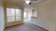 This single-story space showcases a thoughtfully designed open floor plan that connects the living area to the kitchen. The living room is finished with plush, neutral-toned carpeting that adds warmth underfoot, while crisp crown molding runs along the ceiling, lending a polished, refined character to the space. A ceiling fan with a light fixture is centrally positioned, providing both ambient lighting and air circulation.

Natural light filters generously into the living area through large windows dressed with horizontal blinds, offering both brightness and privacy control. The walls are painted in a soft, warm beige tone that complements the overall neutral palette throughout.

A raised kitchen bar with a granite-style countertop serves as both a functional workspace and a visual divider between the living and kitchen areas. The kitchen features track lighting on the ceiling, enhancing task visibility. A pass-through opening with a mirror element above the counter adds depth and visual interest, making the space feel more expansive. White baseboards and clean wall finishes complete the tidy, move-in-ready aesthetic of this well-maintained space.