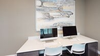 This single-floor business center is a thoughtfully designed workspace that balances functionality with refined aesthetics. The room is anchored by a long, marble-topped desk along one wall, equipped with two Apple iMac computers, wireless keyboards, and mice — offering a clean, modern workstation setup suitable for productivity tasks. Modern white task chairs accompany the desk, providing comfortable seating for users.

A standout feature of the space is the large-scale abstract painting mounted above the workstation, depicting sweeping cloud formations in muted blues, grays, and warm neutrals. The artwork lends the room an artistic, sophisticated character that elevates the otherwise utilitarian purpose of the space.

Dark hardwood-style flooring runs throughout, providing a warm contrast to the neutral taupe walls and light-toned furnishings. A cream-colored lounge chair with an integrated side table offers a secondary seating area, ideal for reading or waiting. A white plantation shutter window allows for adjustable natural light, while a drop ceiling with panel tiles completes the room overhead. A posted "Business Center Rules" sign near the entrance indicates this is a shared or managed facility.