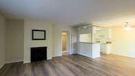 This single-floor space presents an open and functional layout centered around a spacious living area finished with rich hardwood flooring throughout. A sleek, black fireplace anchors one wall of the living room, accompanied by a framed mirror above, adding a classic touch to the otherwise clean, neutral aesthetic. Soft cream-colored walls reflect light evenly, contributing to the overall bright and airy ambiance of the space.

A defining architectural feature is the pass-through counter that connects the living area to the kitchen, framed by a structural beam overhead with decorative corbels. This design element maintains an open sightline between the two areas while subtly defining each zone. The kitchen is outfitted with white cabinetry, a light stone countertop, and white appliances, delivering a clean and practical cooking space.

A doorway visible from the main living area leads to what appears to be a bathroom or hallway, featuring a louvered door detail that adds a subtle textural element. The consistent flooring and cohesive color palette throughout the space create a unified, well-maintained interior that feels both comfortable and move-in ready.
