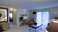 This single-floor space features a well-appointed living area that serves as the focal point of the layout. The room is finished with wall-to-wall carpeting in a neutral tone, contributing to a comfortable and cohesive atmosphere. White walls and a smooth ceiling keep the space feeling open and airy.

A standout feature is the large sliding glass door fitted with vertical blinds, which allows abundant natural light to fill the room while providing access to what appears to be an outdoor area. An air conditioning unit is mounted above the sliding door, ensuring year-round climate control.

The living area comfortably accommodates a cushioned armchair and a decorative wrought-iron console table, which displays framed artwork and accent pieces, adding a touch of character to the space. A matching wrought-iron coffee table anchors the seating area, complementing the overall aesthetic.

A bifold closet door is visible along one wall, offering convenient built-in storage. The overall ambiance of the space is clean, bright, and understated, making it a functional and versatile living environment.