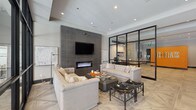 The Flats presents a thoughtfully designed single-story communal space that balances contemporary aesthetics with functional elegance. The entry lobby immediately establishes a bold identity through a vivid orange branded accent wall bearing the property name, complemented by geometric-patterned tile flooring in soft neutral tones. Recessed lighting throughout maintains a clean, polished ceiling plane, while a sculptural multi-bulb chandelier adds an artistic focal point near the entrance. Floor-to-ceiling steel-framed glass partitions divide the space, creating a private office or leasing area that maintains visual connectivity with the main lobby without sacrificing separation.

The lounge area is anchored by a large-format dark tile feature wall housing a sleek linear fireplace and wall-mounted television. Two tufted, neutral-toned sofas flank a substantial dark coffee table, creating a sophisticated and inviting seating arrangement centered on a textured area rug. Black accent chairs and side stools introduce contrast and visual interest. A beverage station with a wine refrigerator and shelving is tucked near the elevator, adding a convenient amenity to the space. The overall ambiance is modern and refined, with a cohesive neutral palette punctuated by bold design accents throughout.