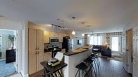 This single-floor space showcases an open-concept layout that integrates the kitchen, dining, and living areas into one cohesive environment. The kitchen is outfitted with stainless steel appliances, including a refrigerator, microwave, and range, complemented by light wood cabinetry that provides ample storage. Dark countertops offer a striking contrast to the cabinetry, and track lighting overhead ensures the workspace is well-illuminated.

A curved breakfast bar serves as the natural dividing point between the kitchen and the rest of the living space, accompanied by several bar-height stools that make it an ideal spot for casual dining or entertaining. Pendant lights hang above the bar, adding a warm, focused glow to the area.

The adjoining living space features hardwood flooring and a distinctly decorated accent wall with a bold, patterned wallcovering that adds visual interest. A ceiling fan with a light fixture provides both airflow and ambient lighting. Natural light filters in through windows at the far end of the room, brightening the space. The overall ambiance is modern and relaxed, with a functional layout designed for everyday living.