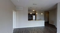 This single-floor space showcases a thoughtfully designed open-concept layout that seamlessly connects the living area to the kitchen. The kitchen is a standout feature, outfitted with dark espresso cabinetry that contrasts elegantly against light granite countertops. A stainless steel refrigerator anchors the cooking area, while a mosaic tile backsplash adds texture and visual interest behind the range. Three pendant lights hang above the peninsula-style breakfast bar, providing both functional task lighting and a decorative focal point that bridges the kitchen and living spaces. The peninsula itself offers additional prep and dining surface space, making it ideal for casual meals or entertaining. Neutral gray walls throughout lend a clean, modern aesthetic that complements the rich, dark hardwood-style flooring running across the entire level. The open floor plan creates a generous sense of space, enhanced by standard ceilings with recessed HVAC venting for efficient climate control. A bi-fold door is visible to one side, likely concealing a closet or utility area. The overall design palette—dark floors, neutral walls, and dark cabinetry—creates a cohesive, contemporary ambiance.