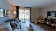 This single-story residence offers a thoughtfully designed layout that balances comfort and functionality across its main living areas. The living room serves as the heart of the home, featuring wood-style flooring, a textured ceiling, and a large sliding glass door framed by dark curtain panels that opens to an outdoor view of greenery, filling the space with natural light. The room accommodates both a seating and media area with ease.

Adjacent to the living room, the dining area is defined by wood-style flooring and a track lighting fixture overhead, with a window allowing additional natural light. An arched passageway connects this space seamlessly to the kitchen, which is equipped with white cabinetry, granite-style countertops, a stainless steel sink, and a striking linear mosaic tile backsplash in shades of blue, black, and grey.

The two bedrooms are carpeted for comfort. The primary bedroom includes two nightstands, warm ambient lighting, and a walk-in closet with built-in shelving. The secondary bedroom features a window with blinds and ample space for a dresser. The bathroom includes wood-style flooring, a granite-topped vanity, and a tub-shower combination.