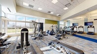 This well-appointed fitness center in Parma, Ohio offers a generous, open floor plan designed to accommodate a wide range of exercise activities. The space is bathed in natural light through large windows that line the walls, providing outdoor views of the surrounding grounds and creating a bright, energizing atmosphere throughout.

The facility is equipped with an extensive selection of cardio machines, including a row of treadmills and elliptical trainers, all positioned to take advantage of the natural light and outdoor views. A dedicated strength training area features multi-functional cable machines, weight benches, and a full dumbbell rack, offering users a complete workout experience.

The interior design is clean and contemporary, with a drop-panel ceiling fitted with both recessed lighting and linear fluorescent fixtures that ensure excellent illumination across the entire space. Patterned, multi-tone carpet tiles in neutral tones with blue accent strips define workout zones throughout the room, while soft teal and sage green accent walls add a refreshing, calming aesthetic.

Wall-mounted flat-screen televisions provide entertainment for cardio users. Glass-paneled doors connect the fitness center to an adjacent lounge or common area, enhancing the sense of openness. The space also includes direct exterior access through a set of glass doors, offering convenient entry and exit.