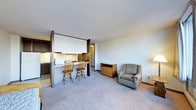 This single-floor space offers an efficient, open-concept layout that combines the living and kitchen areas into one cohesive environment. The kitchen is partially enclosed by a half-wall partition, creating a defined cooking zone while maintaining visual connectivity with the rest of the space. A raised breakfast bar with two wooden bar stools provides a casual dining option, making the space both practical and social.

The kitchen is equipped with essential appliances, including a microwave and stovetop, with upper cabinetry and under-cabinet lighting offering functional storage and workspace illumination. A short corridor visible beyond the kitchen partition suggests access to additional areas of the unit.

The living area is carpeted in a neutral tone, contributing to a warm and understated ambiance. A cushioned armchair and a wooden storage cabinet are positioned along the far wall, providing comfortable seating and storage solutions. A floor lamp nearby adds supplemental lighting to the space.

The textured white ceiling and neutral wall tones throughout create a clean, simple aesthetic. Overall, the space is designed with practicality in mind, offering a straightforward and functional layout suited for everyday living.