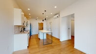 This single-floor unit showcases a thoughtfully designed, modern interior with a clean aesthetic throughout. The open-concept kitchen and living area serves as the heart of the space, featuring warm bamboo hardwood flooring that extends seamlessly through each room. The kitchen is well-appointed with crisp white shaker cabinetry, a white tile backsplash, and a full suite of stainless steel appliances, including a refrigerator, electric range, built-in microwave, and dishwasher. A central island with a white countertop and sleek metal legs provides additional workspace and casual seating, while three pendant lights hang above, adding a modern touch. Recessed lighting and a wall-mounted mini-split HVAC unit contribute to both comfort and a streamlined look. The entry area features a warm wood door flanked by interior closet doors, offering practical storage. The bedroom is bright and airy, highlighted by a large black-framed floor-to-ceiling window that draws in natural light and frames outdoor views, complemented by a sliding barn door for added character. The bathroom maintains the minimalist theme with large-format light tile flooring, a walk-in shower, a wood-toned vanity with a white countertop, and clean white walls throughout.