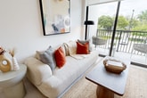 Laguna Gardens' Model 1/1 is a thoughtfully designed one-bedroom, one-bathroom apartment spanning 530 square feet across a single floor. The unit's contemporary aesthetic is defined by crisp white walls, light-toned flooring, and clean architectural lines throughout. The open-concept living and dining area is anchored by large floor-to-ceiling sliding glass doors that flood the space with natural light and provide direct access to a private balcony overlooking lush greenery and palm trees. The kitchen features stainless steel appliances, including a French door refrigerator, electric range, over-the-range microwave, and dishwasher, complemented by two-tone cabinetry with white upper cabinets and wood-grain lower cabinets, white subway tile backsplash, and light quartz countertops. A dining area with a round table sits adjacent to the kitchen. The bedroom offers a serene retreat with floor-to-ceiling windows, ample natural light, and neutral tones. A second bedroom configuration is also visible with warm wood dresser accents and an arched full-length mirror. The bathroom is finished with large-format neutral tiles, a shower-tub combo, and modern fixtures. The unit's entryway features a bold navy door, adding a distinctive design touch to this well-appointed residence.