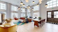 Located at 815 16th Ave N in Nashville, TN, this single-floor community lounge at Scottie is a thoughtfully designed social hub that blends modern aesthetics with vibrant personality. The space is defined by dramatic double-height ceilings that amplify the sense of openness, paired with floor-to-ceiling gridded windows dressed in sheer white curtains that flood the interior with generous natural light. Wide-plank light hardwood flooring runs throughout, lending warmth and continuity to the entire layout. A striking cluster of spherical pendant lights anchors the main lounge area, where plush seating arrangements — including a neutral sofa adorned with rust-toned accent pillows, leather club chairs, and a central wooden dining table with upholstered chairs — create distinct zones for relaxing and gathering. A standout feature is the textured brick accent wall showcasing a lush, layered living moss installation bearing the bold "Beam Me Up Scottie" signage, paired with a solid wood shuffleboard table for entertainment. Sculptural dried-leaf wall art adds an organic, artistic touch above a concierge-style service counter with gold mailboxes nearby. The overall ambiance is energetic yet welcoming, balancing contemporary design elements with curated décor that makes a lasting impression.