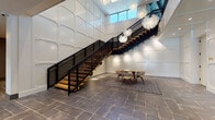 Located at 87 Union St in New Haven, CT, this two-floor amenity space blends contemporary design with warm, natural materials to create a refined and functional environment. The entry level greets visitors with a soaring double-height atrium showcasing a striking open-tread staircase with a sleek black metal railing and wood steps, dramatically lit by cascading sculptural globe pendant lights. White paneled walls and large-format stone tile flooring anchor the space with an air of understated elegance. Glass-partitioned rooms with black steel frames line the corridors, housing intimate co-working and lounge areas furnished with mixed seating arrangements. A sun-drenched sitting area with floor-to-ceiling windows overlooks an outdoor courtyard, flooding the interior with natural light. The kitchen is a standout feature, complete with a large dark island with white countertops, a wood-accented backseat wall with teal LED accent lighting, bar seating, and professional-grade appliances beneath a reclaimed wood ceiling. A spacious multipurpose room with bold teal patterned carpeting accommodates group gatherings, featuring a wall-mounted display, billiards table, and additional seating. Exposed ductwork, warm wood finishes, and wide-plank hardwood floors throughout the second level add an industrial yet inviting character to the overall space.
