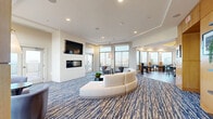 Located at 1855 Saint Francis Street in Reston, VA, this well-appointed amenity lounge offers a generous, open layout across a single floor. The space is defined by an abundance of natural light streaming through large floor-to-ceiling windows and glass doors, framing sweeping panoramic views of the Reston skyline and beyond. A signature blue and cream patterned carpet anchors the main living areas, lending a modern, cohesive aesthetic throughout.

The lounge is thoughtfully divided into distinct functional zones. A relaxed seating area centers around a curved, circular sofa and a wall-mounted flat-screen television above a sleek linear fireplace, creating an inviting focal point for gathering. Adjacent to this is an intimate reading or conversation nook with accent chairs, pendant lighting, and access to an outdoor terrace. Warm wood-paneled accents and built-in shelving add texture and character to the transitions between spaces.

The game room is well-equipped with a full-size billiards table and a shuffleboard table, complemented by casual seating and direct access to the terrace through glass doors. A curved dining area features high-top tables and bar seating set against a sweeping bank of windows, ideal for casual dining or socializing.

The compact kitchenette is finished with marble countertops, a stainless steel sink, and built-in appliances, providing practical support for events and gatherings. A connecting hallway ties the entire space together seamlessly.