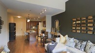 This single-floor residence showcases a thoughtfully designed open-concept layout that seamlessly connects the kitchen, dining, and living areas into one cohesive, flowing space. Rich hardwood flooring extends throughout, grounding the interior with warmth and continuity.

The kitchen is a standout feature, outfitted with dark wood cabinetry, stainless steel appliances, and a central island with bar seating, illuminated by elegant pendant lighting. Track lighting along the ceiling adds further functionality and visual interest to the cooking and dining zones.

A dedicated dining area sits adjacent to the kitchen, furnished with a glass-top table and woven chairs, creating an intimate yet open setting. Crown molding adds a refined architectural detail to the transition between spaces.

The living area anchors the space with a dramatic dark charcoal accent wall, providing a bold contrast to the lighter tones throughout. A comfortable sectional sofa, decorative mirror, and rustic wood wall art contribute to the space's balanced blend of contemporary and warm design elements. The overall ambiance is modern, inviting, and well-appointed.
