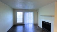 This single-floor residence offers a clean, functional layout designed for comfortable everyday living. The living area serves as the centerpiece of the home, showcasing rich, dark hardwood-style flooring that contrasts elegantly with the light-toned walls. A classic fireplace with a white mantel anchors one side of the room, providing a traditional focal point and a cozy atmosphere. On the opposite wall, a sliding glass door fitted with vertical blinds allows for abundant natural light and offers access to the outdoors, while a side window further brightens the space. The textured ceiling adds a subtle architectural detail consistent throughout the room. The open feel of the living area creates a welcoming and versatile environment, suitable for a variety of furniture arrangements. The neutral color palette of soft whites and warm beige tones throughout the walls provides a blank canvas that complements a wide range of interior styles. Overall, the space presents a practical and inviting single-level layout with key features that enhance both its functionality and aesthetic appeal.