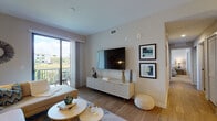 This contemporary single-floor residence offers a thoughtfully designed open-concept layout that seamlessly connects the main living areas. The living room is bright and inviting, featuring wood-look flooring, a wall-mounted flat-screen TV above a sleek white media console, and a sliding glass door that opens to a private balcony with scenic views of lush greenery and the surrounding community. The adjacent dining area is anchored by a warm wood dining table with upholstered seating beneath a statement pendant light, with a large mirror reflecting the kitchen beyond. The kitchen is well-equipped with rich dark cabinetry, stainless steel appliances including a refrigerator, range, and microwave, granite countertops, and a peninsula with an undermount sink that provides additional prep and casual dining space. Two bedrooms offer plush carpeting, generous natural light through horizontal-blind windows, and ceiling fans. The primary bathroom features a frameless glass walk-in shower, tile flooring, a vanity with dark cabinetry, and an adjacent walk-in closet with wire shelving. A secondary bathroom includes a dark wood vanity with a white countertop and tile flooring. Throughout, a neutral color palette and modern finishes create a cohesive, contemporary ambiance.