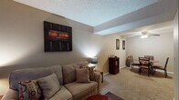 This single-floor residence located at 1900 S Rock Rd in Wichita, Kansas, offers a functional and cohesive layout across several well-defined living spaces. The living room serves as the heart of the home, featuring warm neutral-toned walls, plush carpeting, and a textured ceiling. The space is well-lit and accommodates comfortable seating, complemented by decorative wall art that adds character to the room. Adjacent to the living area, the dining space is equipped with a ceiling fan with light fixture and provides an intimate setting with a round dining table and chairs, framed artwork on the walls, and matching carpeted flooring that ties the spaces together. The entryway offers a welcoming transition into the home, featuring large vertical blinds on a prominent window that allows natural light to filter in. The kitchen is visible from the main living area and features white cabinetry and standard appliances, providing practical functionality. The bedroom is a cozy retreat with carpeted floors, a window with layered curtains for light control and privacy, and ample space for furnishings. The full bathroom includes a tub-shower combination with chrome fixtures, tile flooring, and a vanity with mirror. Throughout the residence, the consistent warm beige color palette and carpeted floors create a cohesive and comfortable atmosphere.