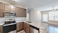 This single-floor residence offers a thoughtfully designed layout across two bedrooms and two bathrooms. The open-concept kitchen serves as a central hub of the home, featuring rich medium-toned wood cabinetry, granite-look countertops, a white subway tile backsplash, stainless steel appliances including a refrigerator, range, and over-the-range microwave, and a peninsula with a double sink that overlooks the adjoining living area. The kitchen flows seamlessly into a spacious living room finished with durable wood-style plank flooring, a ceiling fan with light fixture, and a glass door providing access to an outdoor balcony area with natural light.

Both bedrooms are carpeted and feature large windows that allow ample natural light and exterior views of the surrounding neighborhood. The primary bedroom includes access to a dedicated walk-in closet outfitted with multi-tiered wire shelving for organized storage. The bathrooms mirror the home's design aesthetic with matching wood cabinetry, granite-look countertops, and wood-style flooring. Neutral gray-beige tones are carried throughout, creating a cohesive and modern ambiance. A hallway connects the main living spaces to the bedrooms and bathrooms.