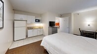 Located in Siegel, Bartlett, Tennessee, this self-contained studio room offers a practical and comfortable layout designed for extended stays. The main living area features a queen-sized bed dressed in crisp white bedding, complemented by a upholstered headboard and wall-mounted lighting on either side. Warm, neutral-toned walls and patterned carpeting contribute to a welcoming ambiance throughout the sleeping area. A window with drape curtains allows natural light to filter in, while a wall-mounted HVAC unit ensures year-round climate control. A flat-screen TV atop a multi-drawer dresser provides in-room entertainment, and a small table with seating offers a dedicated dining or work area. The kitchenette is well-equipped with white cabinetry, a granite-style countertop, a microwave, a full-size refrigerator, and a sink — ideal for preparing meals during longer stays. Tiled flooring in the kitchenette transitions seamlessly into the adjacent bathroom, which features a granite vanity countertop, a large mirror, tile floors, and accessibility-friendly grab bars. The shower area is enclosed with a brightly colored curtain, adding a pop of contrast to the otherwise neutral palette of the room.