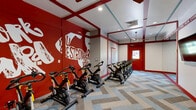 Located in Lenexa, Kansas, this dedicated spin studio is a purpose-built exercise room designed to inspire and motivate. The space makes an immediate visual impact with its striking red-and-white color scheme, carried through bold trim work, red-framed doors, and energetic motivational murals painted directly on the walls — featuring phrases such as "Work Hard," "Be Stronger," and "You Are Stronger Than You Thought You Could Be." A row of professional-grade stationary cycling bikes lines the room, providing ample capacity for group spin sessions. The floor features a distinctive herringbone-patterned carpet in muted tones of blue, gray, and tan, adding a dynamic visual texture that contrasts well with the bold wall décor. Recessed ceiling lighting keeps the room well-lit, while a ceiling fan ensures comfortable airflow during intense workouts. Red accent lines extend across the ceiling, reinforcing the energetic design language throughout. Large mirrored panels on one wall visually expand the space and allow participants to monitor their form. Multiple entry doors with windowed panels allow for natural light and easy access. The overall ambiance is high-energy and motivational, making this studio an ideal environment for focused, group fitness training.