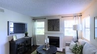 This single-floor living space centers around a white-surround fireplace, which serves as the room's focal point and is accented by a decorative tree-motif wall panel mounted above. The living area features dark hardwood flooring that contrasts warmly with the light neutral wall tones, creating a balanced and inviting atmosphere. A textured popcorn ceiling adds a classic architectural detail overhead. Two windows dressed with sheer white curtains and dark curtain rods allow generous natural light to fill the room, offering views of the surrounding greenery outside. A dark wood media console positioned along one wall houses a flat-screen television, complemented by small decorative accents including a potted plant. The seating area incorporates a plush sofa with green patterned throw pillows, arranged around a round dark wood coffee table styled with candle holders and books. A green area rug ties the seating arrangement together, adding texture and warmth to the space. Overall, the room presents a comfortable and well-appointed interior with a blend of rustic and traditional design elements.