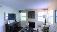 This single-floor living space centers around a white-surround fireplace, which serves as the room's focal point and is accented by a decorative tree-motif wall panel mounted above. The living area features dark hardwood flooring that contrasts warmly with the light neutral wall tones, creating a balanced and inviting atmosphere. A textured popcorn ceiling adds a classic architectural detail overhead. Two windows dressed with sheer white curtains and dark curtain rods allow generous natural light to fill the room, offering views of the surrounding greenery outside. A dark wood media console positioned along one wall houses a flat-screen television, complemented by small decorative accents including a potted plant. The seating area incorporates a plush sofa with green patterned throw pillows, arranged around a round dark wood coffee table styled with candle holders and books. A green area rug ties the seating arrangement together, adding texture and warmth to the space. Overall, the room presents a comfortable and well-appointed interior with a blend of rustic and traditional design elements.