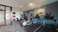 Located at 1224 West Riverside Avenue in Spokane, Washington, this well-appointed fitness center offers a comprehensive range of exercise equipment and amenities within a thoughtfully designed single-level space. The room features a textured ceiling with track lighting that evenly illuminates the area, while patterned carpet tiles cover the floor throughout.

The cardio zone is equipped with multiple treadmills, elliptical trainers, and stationary bikes — several of which feature interactive display screens. A dedicated strength training area includes a multi-function cable machine, a full dumbbell rack ranging across various weights, an adjustable workout bench, and an assortment of exercise balls in multiple sizes for functional fitness.

A wall-mounted television and a yoga mat rack provide additional convenience for users. The space is finished with deep teal accent walls adorned with black-and-white framed photographs, lending a modern and polished aesthetic.

A standout feature of this fitness center is the floor-to-ceiling glass partition and door that opens directly onto an outdoor terrace, offering views of the surrounding Spokane cityscape. The terrace, furnished with seating, provides a refreshing outdoor retreat accessible directly from the gym floor, making this fitness facility both functional and uniquely inviting.