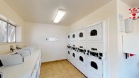 This shared laundry facility is a well-organized, single-floor space designed to accommodate high-volume use efficiently. The room is lined with multiple front-loading washing machines positioned along the walls, complemented by several stacked dryer units that maximize vertical space. The layout is thoughtfully arranged to allow comfortable movement throughout the room while providing access to all machines.

The interior features neutral white walls and durable tile flooring in a warm beige tone, creating a clean and practical environment. Overhead fluorescent lighting ensures the space is well-lit, while horizontal windows along the upper portion of the walls allow natural light to filter in, offering views of the surrounding outdoor greenery. A glass-paned entry door further contributes to the natural brightness of the room.

Functional amenities include folding tables for convenient laundry sorting and folding, a wall-mounted soap dispenser, informational postings, and a prominently displayed fire extinguisher for safety compliance. A bulletin board is also present for resident communication. The overall ambiance is clean, utilitarian, and efficiently designed to serve the practical laundry needs of a multi-unit residential community.