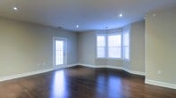 This single-floor space presents a clean, modern aesthetic defined by thoughtful architectural details and quality finishes throughout. The main living area immediately draws attention with its rich, dark hardwood flooring that runs seamlessly across the open layout, reflecting light and adding warmth to the overall ambiance.

Natural light fills the room through a classic bay window arrangement, which creates a welcoming alcove and adds visual depth to the space. A secondary door with a traditional grid-pane window provides additional brightness while offering convenient access. Recessed ceiling lights complement the natural light, ensuring the space remains well-lit at all times.

The walls are finished in a neutral, warm gray tone that pairs harmoniously with the white trim and baseboards, lending the space a crisp and polished look. The clean lines of the trim work throughout reinforce the space's contemporary yet timeless character.

The open floor plan provides generous room to configure the space flexibly, making it well-suited for a variety of uses. The combination of hardwood floors, bay windows, and recessed lighting creates an inviting atmosphere that balances both style and functionality, making this a versatile and appealing space overall.