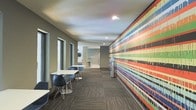 This single-floor space makes an immediate visual impact through its bold, floor-to-ceiling mural that runs the length of one wall, showcasing a dynamic composition of horizontal stripes in vivid reds, blues, greens, yellows, and blacks. The artwork serves as the defining architectural feature of the space, drawing the eye down the corridor and creating a strong sense of energy and movement.

The flooring throughout consists of patterned carpet tiles in a neutral charcoal tone, providing a grounded contrast to the colorful mural. Casual seating areas are arranged along the opposite wall, featuring modern molded chairs in navy blue paired with white rectangular tables, offering functional spots for informal gatherings or work. Natural light filters in through a series of windows positioned along the left wall, complementing the recessed ceiling lighting above.

Toward the far end of the corridor, additional amenities are visible, including what appears to be a small service or utility area. The neutral gray walls and smooth white ceiling keep the overall palette balanced, allowing the mural to remain the focal point. The space conveys a modern, creative atmosphere suited for collaborative or communal use.