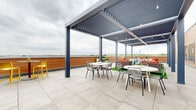 This rooftop terrace is a well-designed outdoor amenity space that combines open-air and semi-covered areas to create a versatile, social environment. The terrace is finished with large-format light gray porcelain tiles throughout, providing a clean, contemporary foundation that extends across the entirety of the space.

A prominent louvered pergola serves as the architectural centerpiece, constructed from steel posts in a dark charcoal finish with adjustable slatted roofing panels. String lights are strung beneath the pergola structure, adding an ambient quality suited for evening use. The pergola spans a generous portion of the terrace, sheltering multiple seating arrangements that include round white dining tables paired with wire-frame chairs, as well as woven rattan lounge furniture accented with orange cushions positioned on artificial turf zones.

The perimeter of the terrace features horizontal wood-tone cladding, which adds warmth against the industrial metal framework. Access to and from an adjacent interior space is provided through multiple dark-framed glass entry doors and windows, allowing visual connectivity between indoors and outdoors. A low wood-paneled parapet wall runs along the outer edge of the rooftop, complementing the skyline views visible beyond. The city skyline is visible in the distance, offering a compelling backdrop to the entire terrace.