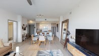 This single-floor residence showcases a thoughtfully designed open-concept layout that connects the living, dining, and kitchen areas into one cohesive, light-filled space. Light-toned hardwood flooring runs throughout, contributing to a clean and airy aesthetic that ties each zone together seamlessly.

The kitchen is outfitted with flat-panel cabinetry in a warm wood finish, stainless steel appliances including a refrigerator and range, and a peninsula that doubles as a breakfast bar with seating. Track lighting overhead provides functional illumination while enhancing the modern feel.

Adjacent to the kitchen, the dining area comfortably accommodates a full glass-top table surrounded by woven wood chairs, making it well-suited for both everyday meals and entertaining. A large leaning mirror nearby adds depth and reflects natural light throughout the space.

The living area is defined by a patterned area rug with blue and white tones, anchoring the seating zone in front of a media console. Decorative accent pieces and a ceiling fan add personality and comfort. White walls and a neutral palette throughout create a bright, contemporary ambiance.
