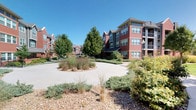 This outdoor amenity space is thoughtfully designed to offer residents a comprehensive recreational experience. At its center is a generously sized swimming pool with crystal-clear water, surrounded by an expansive concrete deck lined with lounge chairs and patio umbrellas, ideal for sunbathing and relaxation. Adjacent to the pool sits a circular hot tub, partially sheltered beneath a white pergola structure supported by decorative brick columns, which provides a shaded transition zone between the various outdoor areas.

The covered patio sections feature comfortable seating arrangements, including cushioned sofas, lounge chairs, and accent tables, creating inviting spaces for both casual conversation and unwinding. Grilling stations are conveniently integrated into the covered areas, enhancing the space's functionality for outdoor entertaining. Colorful accent furniture and ornamental grasses add visual interest throughout.

The architecture of the surrounding multi-story buildings incorporates a combination of brick, painted siding in warm earth tones, and large windows, contributing a residential warmth to the setting. Wrought iron fencing defines the perimeter of the amenity area, while mature trees and manicured landscaping provide natural shade and a pleasant green backdrop. The space benefits from abundant natural light under open skies, making it a vibrant and welcoming outdoor destination.