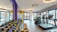 This single-floor fitness center is designed to deliver a comprehensive workout experience within a bright, contemporary space. The layout is thoughtfully organized to accommodate both cardio and strength training needs. Along the window-facing wall, a row of Life Fitness treadmills, ellipticals, and a stair climber are positioned to take full advantage of the large floor-to-ceiling windows, which flood the space with natural light and offer open views of the surrounding streetscape. A dedicated cardio section and freestanding exercise bike round out the aerobic offerings. The strength training area is anchored by a yellow Life Fitness functional cable trainer, complemented by a full rack of dumbbells arranged along a mirrored, purple-accented wall — a bold design choice that adds energy and personality to the space. A flat bench and additional free weights provide flexibility for a variety of exercises. The flooring transitions between durable rubber and smooth surfaces suited for different training zones. Overhead, distinctive circular pendant light fixtures and recessed lighting provide warm, even illumination throughout. A ceiling-mounted HVAC unit ensures climate comfort year-round. The entrance features glass double doors, reinforcing the open, airy aesthetic that defines this well-appointed fitness facility.