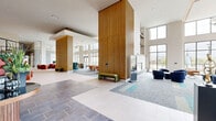 Located at 1133 West Baltimore Pike in Media, Pennsylvania, this impressive single-floor amenity space offers a generously scaled, contemporary environment designed for comfort and community. The grand lobby serves as the centerpiece, boasting soaring double-height ceilings, large floor-to-ceiling windows that flood the interior with natural light, and a sophisticated mix of large-format tile and plush carpet flooring. Striking vertical wood-slat columns add warmth and architectural character throughout the open space.

Multiple seating areas are thoughtfully arranged across the lobby, featuring an eclectic mix of navy blue lounge chairs, bold red armchairs, and teal benches, all accented by curated artwork and lush greenery. A central refreshment bar, outfitted with wood cabinetry, stainless steel appliances, and pendant lighting, provides a convenient amenity for residents and guests. A dramatic wood-tread staircase with black metal railings and integrated step lighting serves as a focal architectural statement, connecting levels in style.

Additional highlights include a well-appointed mailroom, a living green wall, a geometric chandelier, and direct views of the surrounding landscape. The overall ambiance is modern, welcoming, and meticulously designed to foster an engaging community atmosphere.