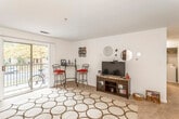 This single-floor residence offers a practical and comfortable layout with distinct living, dining, kitchen, bedroom, and bathroom areas. The living room is spacious, featuring carpet flooring topped with a bold geometric area rug. A sliding glass door fills the room with natural light and opens to a private balcony with outdoor views of surrounding trees. The open sightline from the living room extends into a defined dining area, which accommodates a four-person table and connects visually to the kitchen. The galley-style kitchen is efficiently designed with warm wood cabinetry lining both walls, ample counter space, tile flooring, and white appliances including a range and refrigerator. A passthrough connects the kitchen to the main living and dining spaces. The bedroom is carpeted and generously sized, featuring a large window with natural light, a walk-in closet with built-in shelving, and ample room for a full furniture arrangement. The bathroom includes a single-sink vanity with wood cabinetry, a large wall mirror, and a shower-tub combination. Throughout the unit, neutral wall tones create a cohesive, understated ambiance.