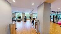 This generously sized fitness center spans a single floor and offers a comprehensive workout environment designed to accommodate a wide range of exercise needs. The space features warm hardwood-style flooring throughout, recessed ceiling lighting, and a modern, neutral color palette of soft gray and white tones that creates a clean, professional atmosphere.

Large floor-to-ceiling windows line the perimeter, flooding the interior with abundant natural light and providing calming views of mature trees and greenery outside. The cardio zone is prominently featured, housing a row of treadmills by True Fitness, multiple elliptical cross-trainers, and a recumbent bike, all positioned to take advantage of the scenic outdoor views.

A dedicated free weights and functional training area is equipped with a barbell rack, adjustable benches, stability balls, a BOSU trainer, and a dumbbell rack. Full-length mirrors cover an entire wall in this section, enhancing the sense of space and supporting proper form during training. A parallel bar and adjustable workout bench provide additional rehabilitation and strength training options.

The space also benefits from a ceiling-mounted HVAC system ensuring year-round climate comfort. The overall layout is open and well-organized, making the fitness center both functional and inviting for users of all fitness levels.