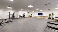 Located at 50 Otterbein Rd, this well-appointed fitness center offers a generous single-floor layout designed to accommodate a full range of exercise activities. The space is finished with clean white walls and a drop ceiling fitted with recessed flat-panel lighting that ensures bright, even illumination throughout. Durable speckled carpet flooring covers the entire area, providing a practical and comfortable surface underfoot.

The exercise room is comprehensively equipped to support diverse fitness routines. A cardio zone features multiple treadmills and an elliptical trainer, each equipped with individual display screens, complemented by wall-mounted flat-screen TVs for entertainment during workouts. A strength training section includes a multi-station cable machine, a weight rack with barbells, a dumbbell rack with a full range of free weights, and an adjustable bench. A stationary cycle adds further cardio variety. Full-length mirrors along one wall help maximize the sense of openness while assisting users with form and technique.

Storage is thoughtfully addressed with a low open shelving unit fitted with wicker baskets along one wall. Tall potted plants near the entrance add a touch of greenery to the otherwise modern, minimalist aesthetic. Double doors provide easy access, and the overall layout leaves ample open floor space at the center, making the room suitable for stretching, bodyweight exercises, or group fitness activities.