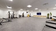 Located at 50 Otterbein Rd, this well-appointed fitness center offers a generous single-floor layout designed to accommodate a full range of exercise activities. The space is finished with clean white walls and a drop ceiling fitted with recessed flat-panel lighting that ensures bright, even illumination throughout. Durable speckled carpet flooring covers the entire area, providing a practical and comfortable surface underfoot.

The exercise room is comprehensively equipped to support diverse fitness routines. A cardio zone features multiple treadmills and an elliptical trainer, each equipped with individual display screens, complemented by wall-mounted flat-screen TVs for entertainment during workouts. A strength training section includes a multi-station cable machine, a weight rack with barbells, a dumbbell rack with a full range of free weights, and an adjustable bench. A stationary cycle adds further cardio variety. Full-length mirrors along one wall help maximize the sense of openness while assisting users with form and technique.

Storage is thoughtfully addressed with a low open shelving unit fitted with wicker baskets along one wall. Tall potted plants near the entrance add a touch of greenery to the otherwise modern, minimalist aesthetic. Double doors provide easy access, and the overall layout leaves ample open floor space at the center, making the room suitable for stretching, bodyweight exercises, or group fitness activities.