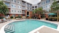 Located at 712 Tucker St in Raleigh, NC, this thoughtfully designed outdoor amenity courtyard offers a comprehensive set of communal features within a landscaped, multi-building complex. The centerpiece is a generously sized resort-style swimming pool with clear turquoise water, surrounded by a paved deck furnished with lounge chairs and umbrellas for relaxed outdoor enjoyment. Adjacent to the pool, a tiled circular hot tub is nestled into a brick-accented surround, providing a spa-like retreat. A dedicated outdoor kitchen area features a built-in stainless steel grill with granite countertops set atop a brick base, ideal for social gatherings. A cozy sunken fire pit lounge, framed by lush greenery and low stone walls, offers seating around a decorative glass-bead fire table. Winding concrete walkways bordered by manicured shrubs and plantings connect the various amenity zones throughout the courtyard. A pergola-shaded pavilion provides additional covered gathering space near the pool. The interior connector corridor features speckled terrazzo flooring, wainscoting, ceiling fans, and vibrant abstract artwork, lending a polished, welcoming transition between the outdoor spaces. The surrounding multi-story brick and contemporary facade buildings frame the courtyard, creating an enclosed, community-oriented atmosphere.