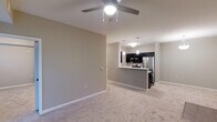 This single-floor space showcases an open-concept layout that seamlessly connects the living area to the kitchen. The living area is finished with plush wall-to-wall carpeting and neutral-toned walls, creating a clean, versatile backdrop. A ceiling fan with an integrated light fixture is centrally mounted, providing both air circulation and ambient lighting.

The kitchen is defined by dark cabinetry that contrasts sharply against the lighter walls and countertops, lending a modern aesthetic. A peninsula-style counter serves as a natural divider between the kitchen and living space, offering additional workspace and the potential for casual seating. Stainless steel appliances, including a refrigerator and microwave, are integrated into the cabinetry, contributing to a sleek, cohesive look. Pendant lights hang above the kitchen area, providing focused task lighting that complements the overall design.

White baseboards frame the perimeter of the space, adding a crisp finishing detail throughout. The open layout and consistent neutral palette create a bright, airy atmosphere, making the space feel welcoming and adaptable to a variety of furnishing styles.