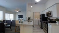 This single-floor residence is designed with an open-concept layout that seamlessly connects the kitchen and living area, creating a spacious and cohesive environment. The kitchen serves as the centerpiece, anchored by a peninsula with a granite-style countertop that provides both prep space and casual seating for two. White shaker cabinetry lines the upper and lower walls, complemented by stainless steel appliances including a range and refrigerator. A pendant light hangs above the sink, adding a focused decorative element to the workspace.

The light hardwood-style flooring extends throughout, unifying the kitchen and living spaces visually. The living area features a ceiling fan with integrated lighting and a wall-mounted flat-screen television, contributing to a practical and comfortable setup. Large windows allow natural light to fill the space, enhancing the bright, airy atmosphere established by the neutral white and soft gray wall tones.

Notably, the unit includes a dedicated in-unit laundry area with a full-size washer and dryer, conveniently accessible directly from the kitchen. The overall design reflects a clean, modern aesthetic with functional finishes suited for everyday living.